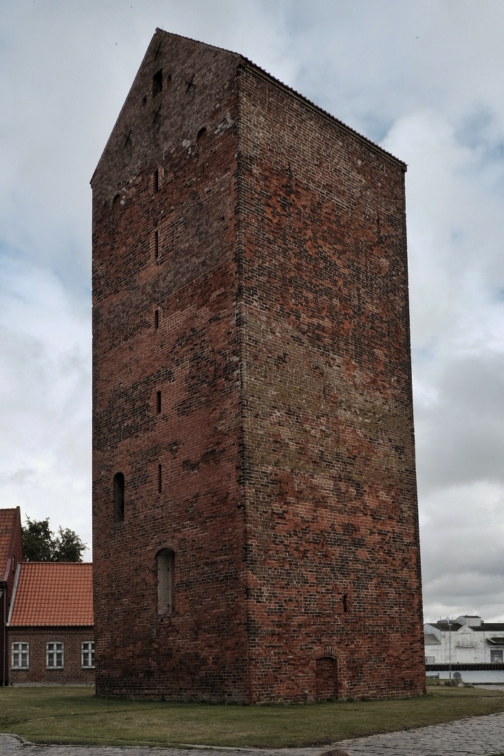 Alter Backsteinturm aus roten Ziegeln mit schmalen Fensteröffnungen, umgeben von Gras und angrenzenden Gebäuden.