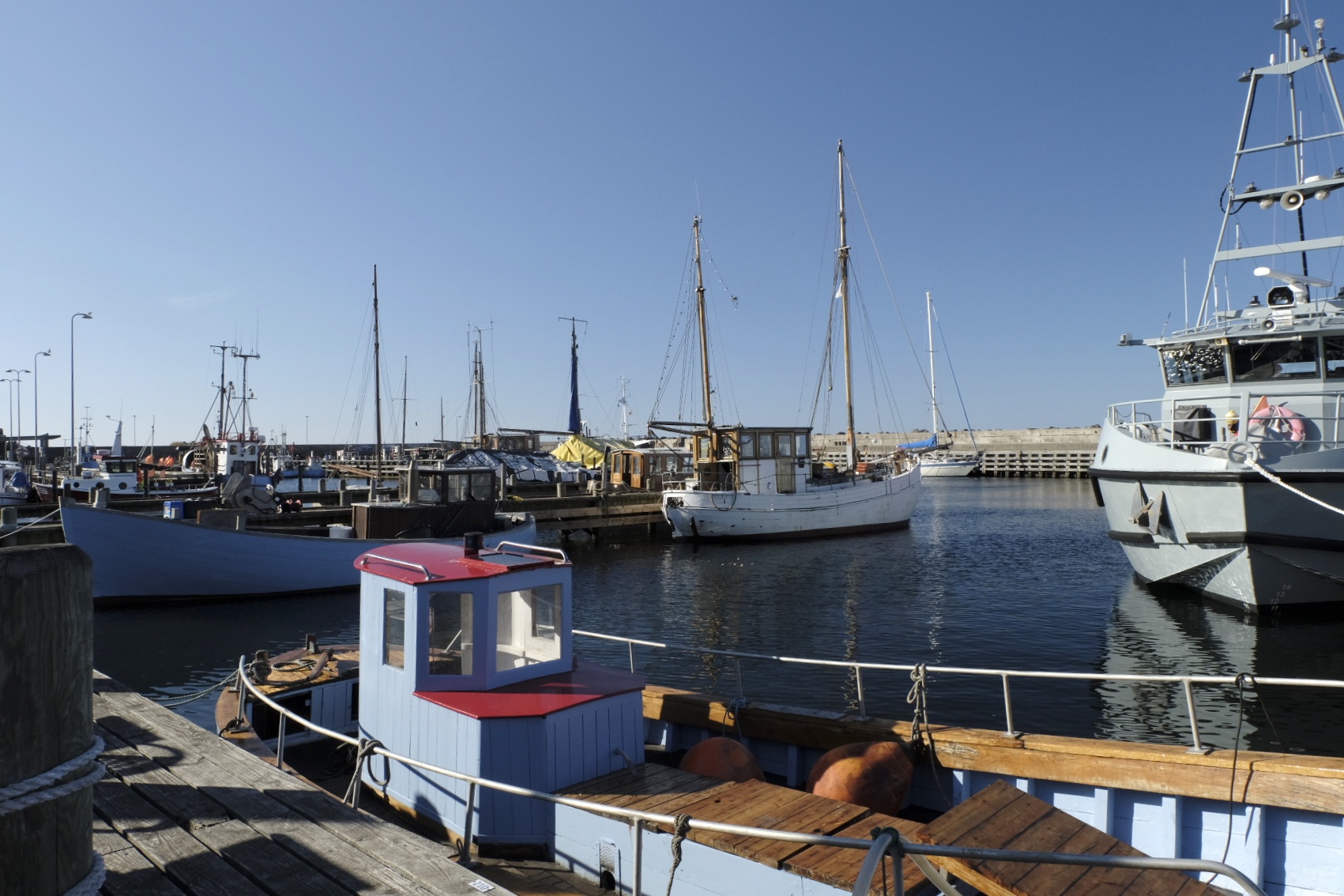 Fischereihafen mit verschiedenen Booten und Segelschiffen, hölzerne Anlegestelle, klarer Himmel.