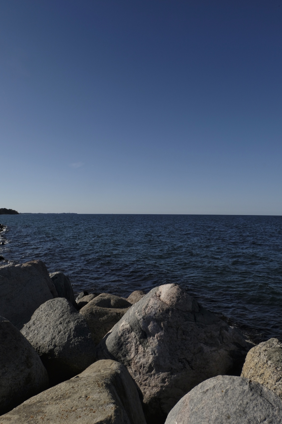 Felsige Küste mit großen Granitsteinen im Vordergrund und ruhigem, blauem Meer unter klarem Himmel.