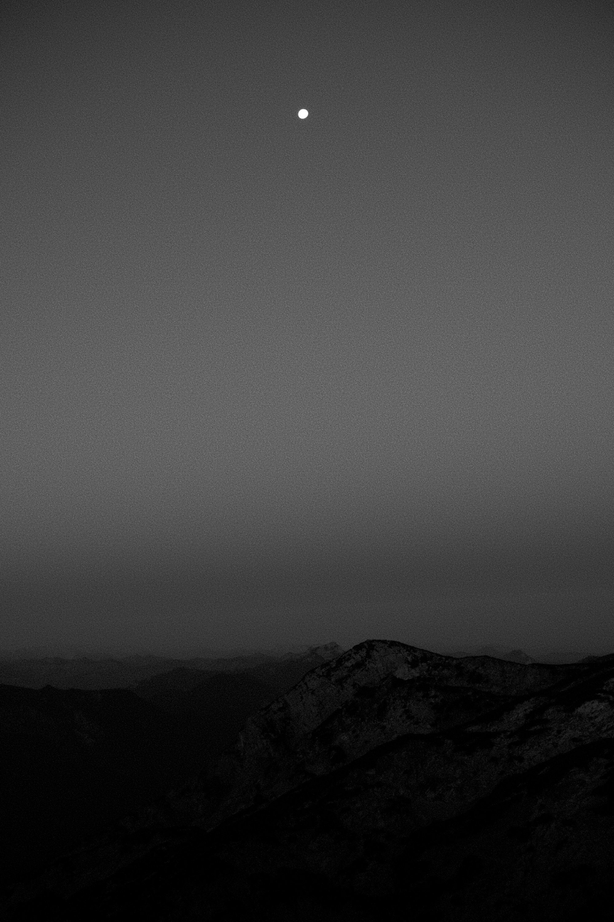 Monochrome Nachtaufnahme: Mond über dunkler Bergsilhouette gegen grauen Himmel.