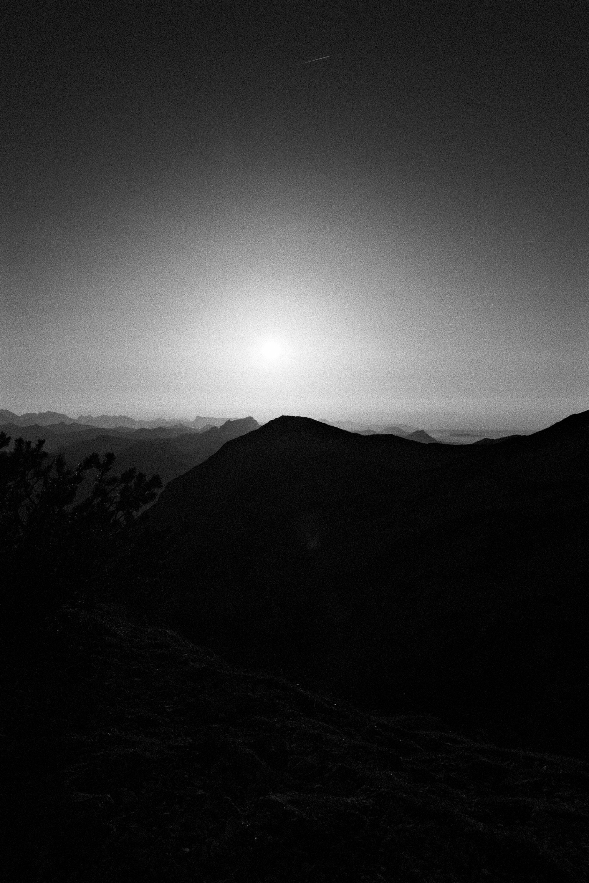 Schwarzweiß-Berglandschaft bei Sonnenaufgang oder -untergang mit silhouettierten Hügeln und hellem Himmel.