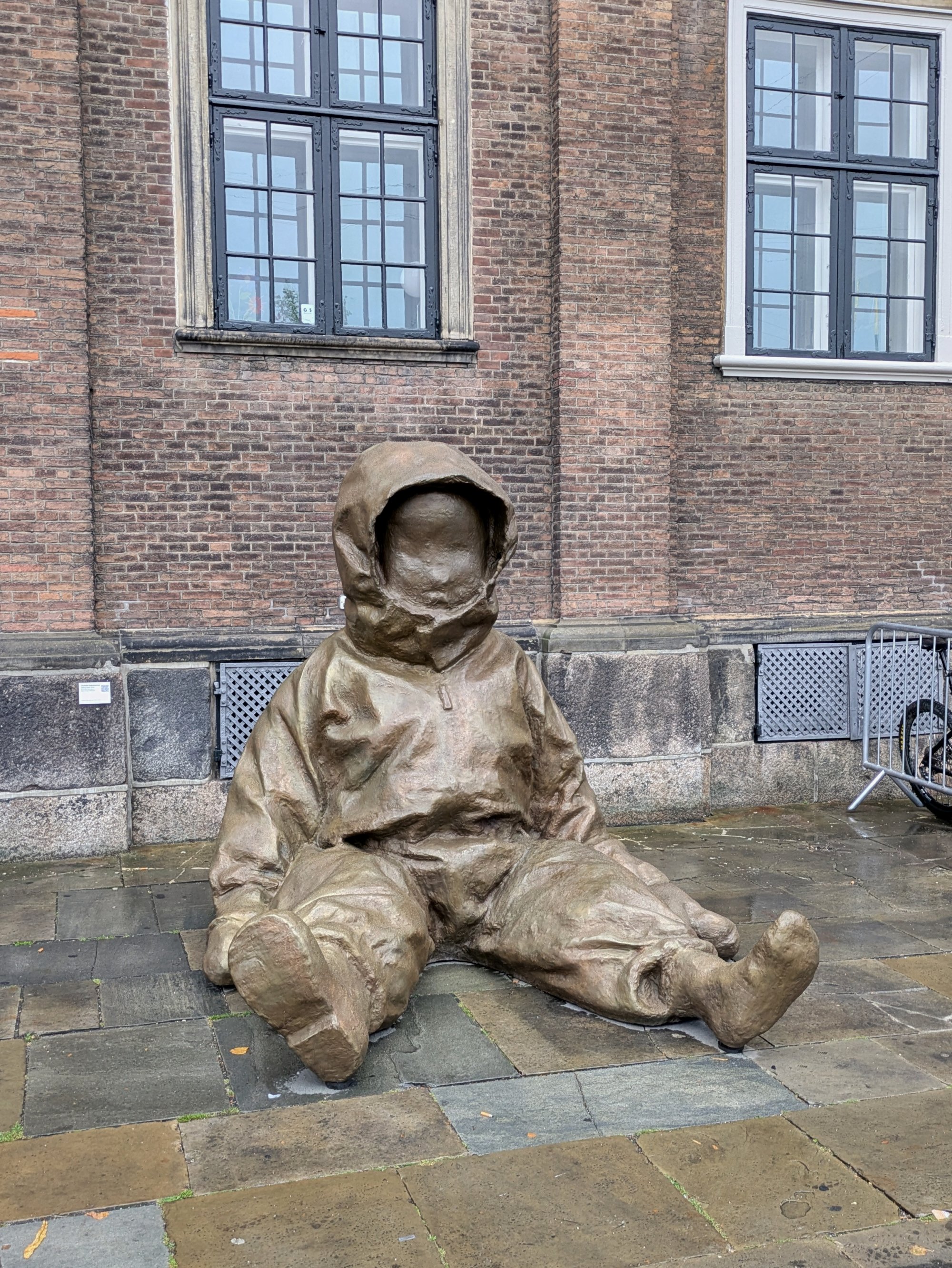 Bronzeskulptur eines sitzenden Kindes mit Kapuze vor Backsteingebäude mit Fenstern.