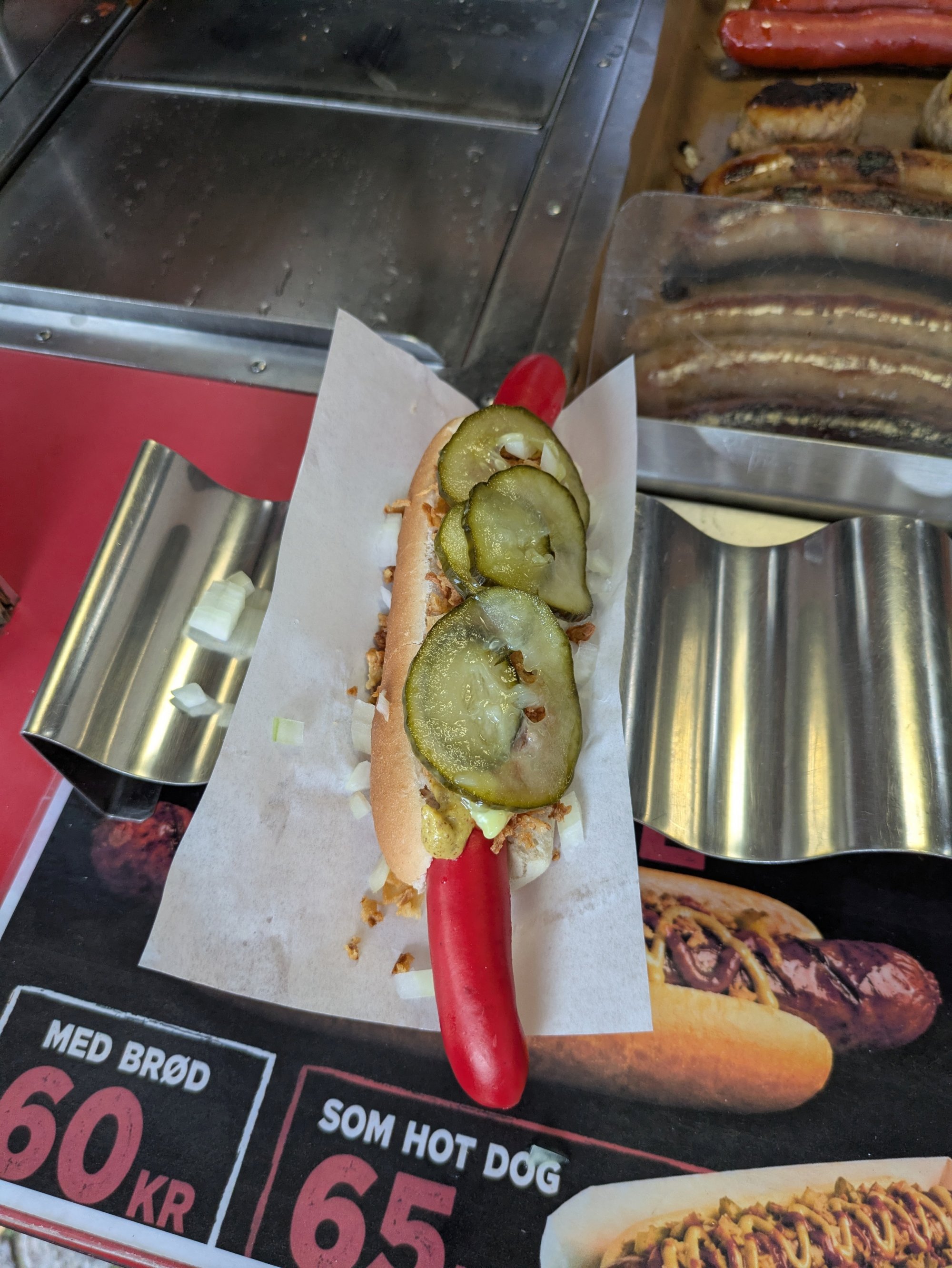 Hot Dog mit Gurken und Zwiebeln auf rotem Holder, im Hintergrund Würstchen in Verkaufsstand.