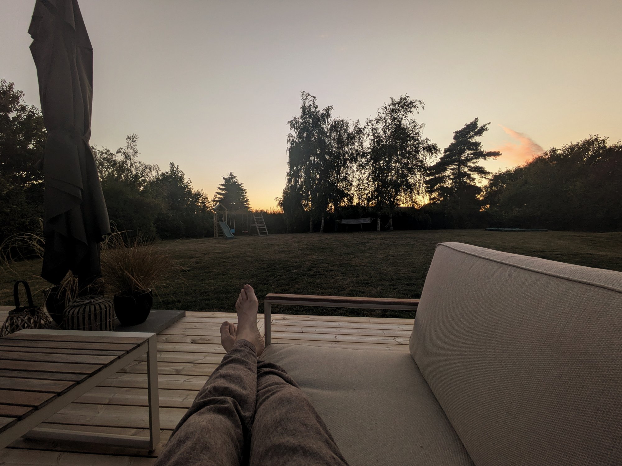 Person sitzt auf Terrasse und blickt bei Sonnenuntergang auf große Wiese mit Bäumen und Spielgeräten.