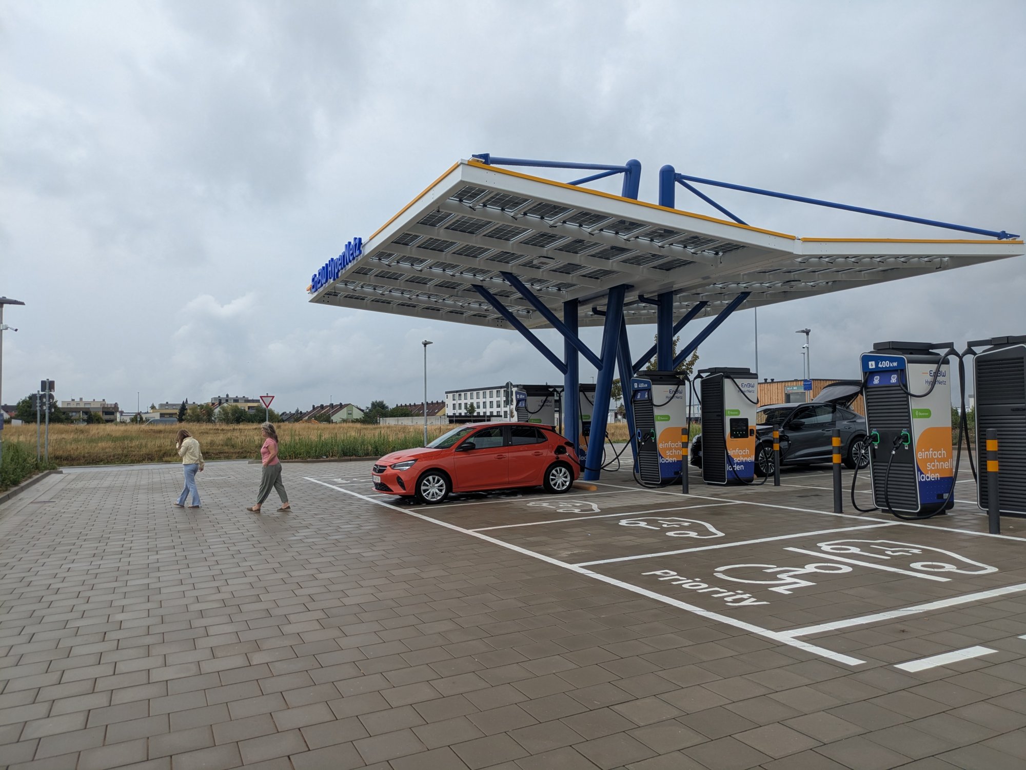 Elektroauto an Schnellladestation unter Solardach, zwei Personen gehen vorbei, ländliche Umgebung.