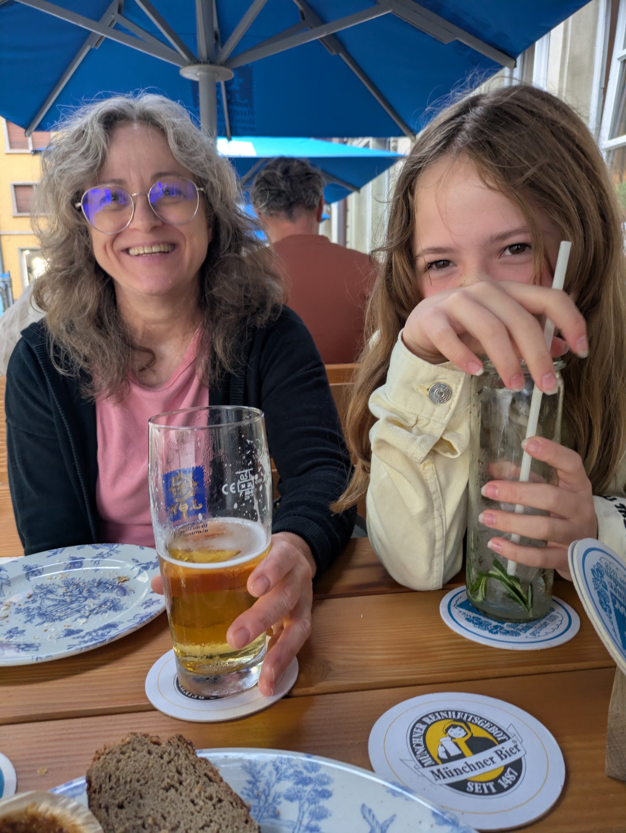 Zwei Personen sitzen unter blauem Sonnenschirm an Tisch mit Bier und Brotzeit.
