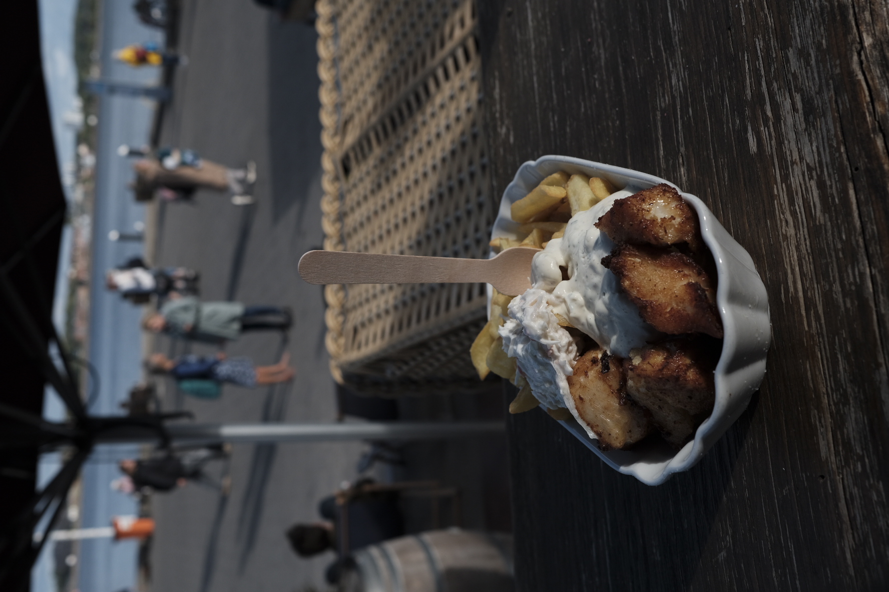 Fischgerichte mit Pommes und Mayonnaise in einer weißen Schale an einem Strandpromenade mit Menschen im Hintergrund.