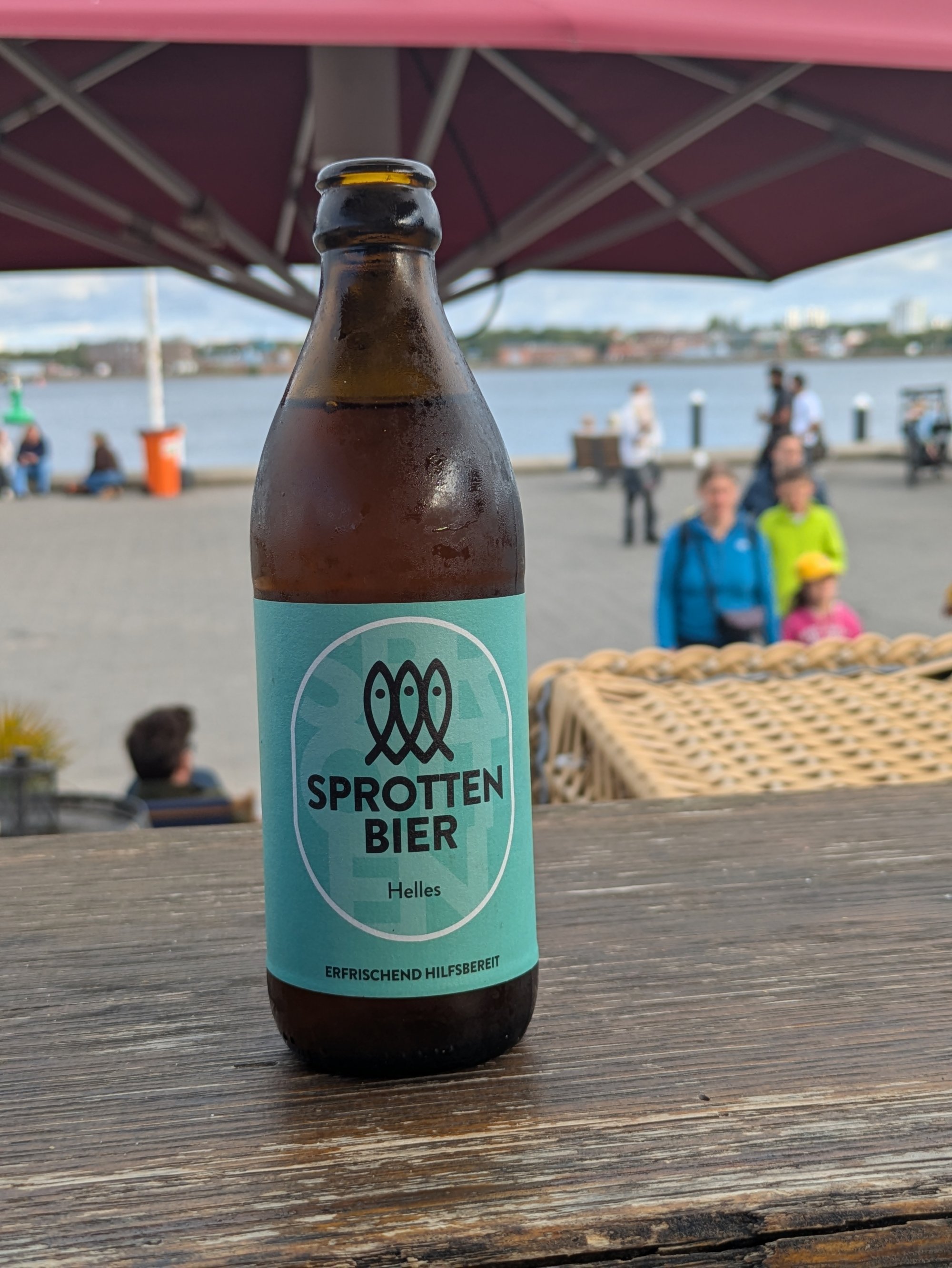Sprotten Bier Flasche auf Holztisch am Hafen mit Schirm, Menschen und Wasser im Hintergrund.