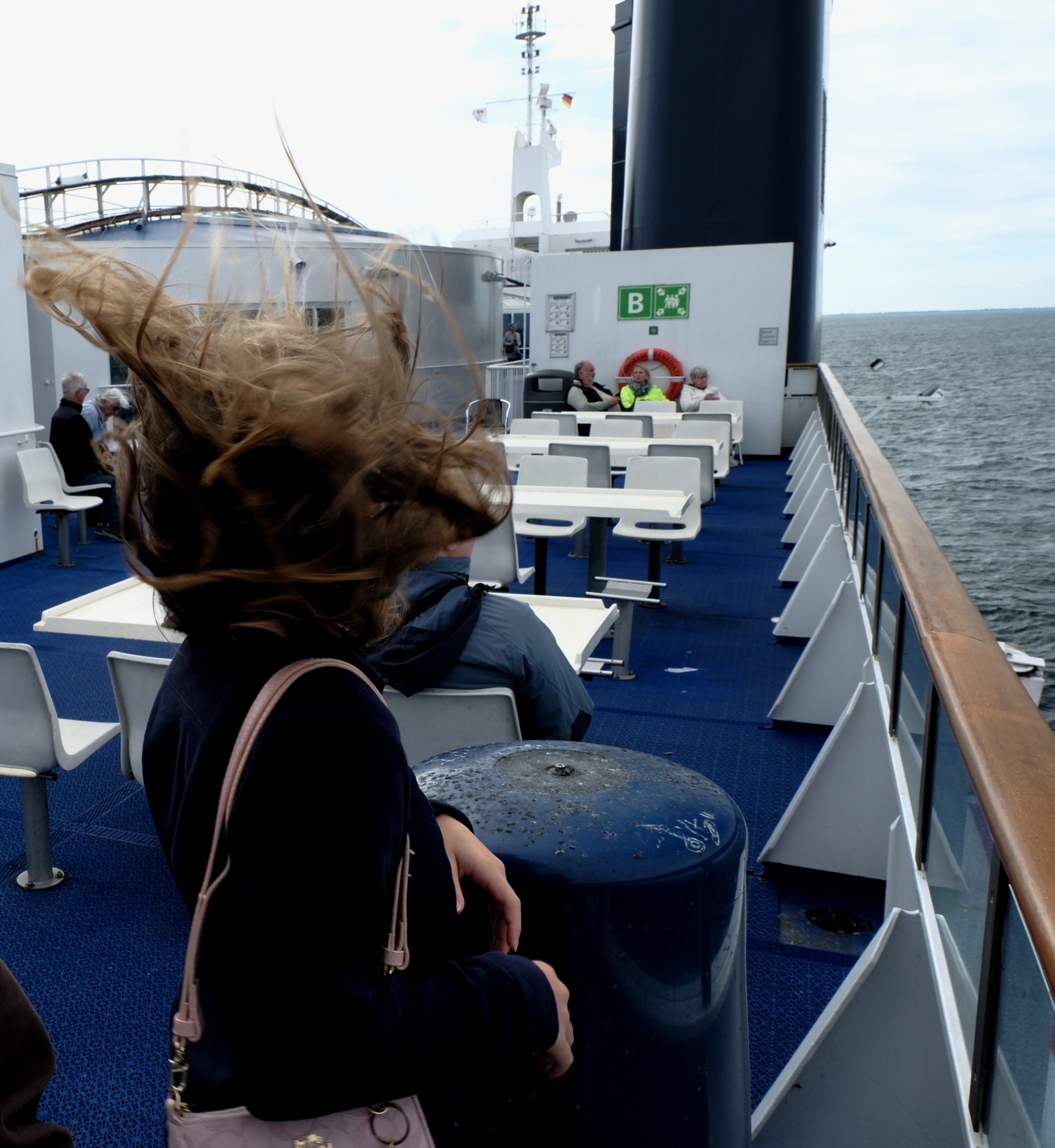 Frau mit wehenden Haaren auf dem Oberdeck einer Fähre, blickt aufs Meer mit anderen Passagieren.